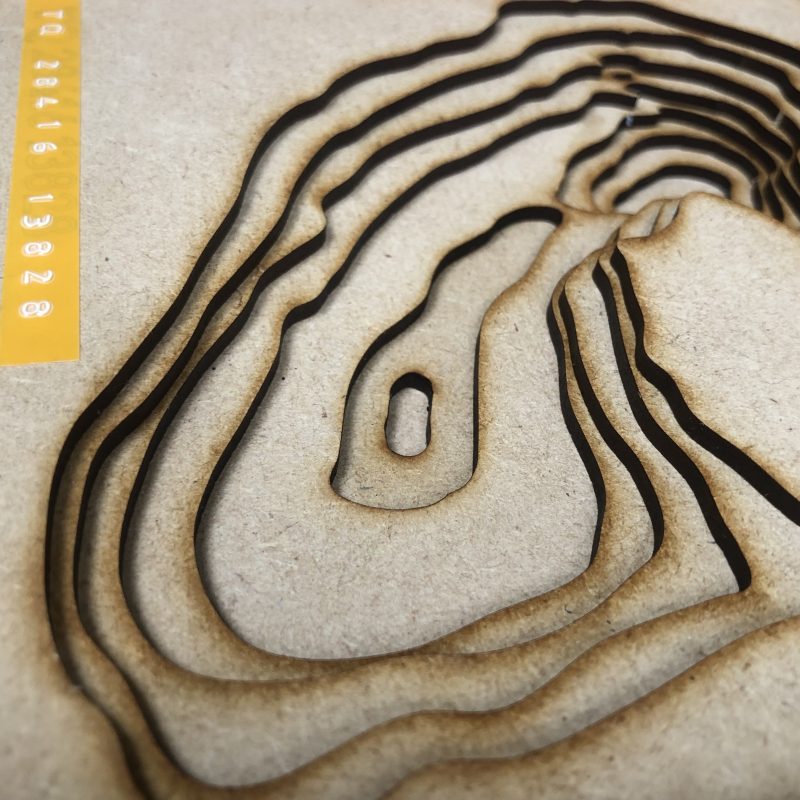 The artwork is a topographic style piece created using layered, laser-cut wood, enclosed in a wooden frame made of upcycled plywood, emphasizing its handcrafted natural aesthetic.