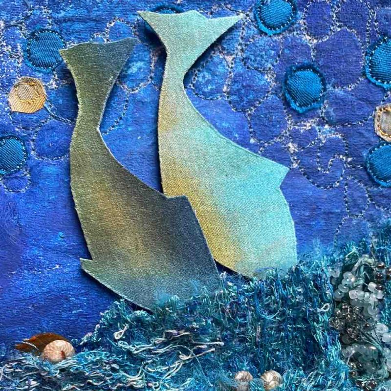 Two fish on a stitched & embroidered background, in shades of blue, embellishe with shells