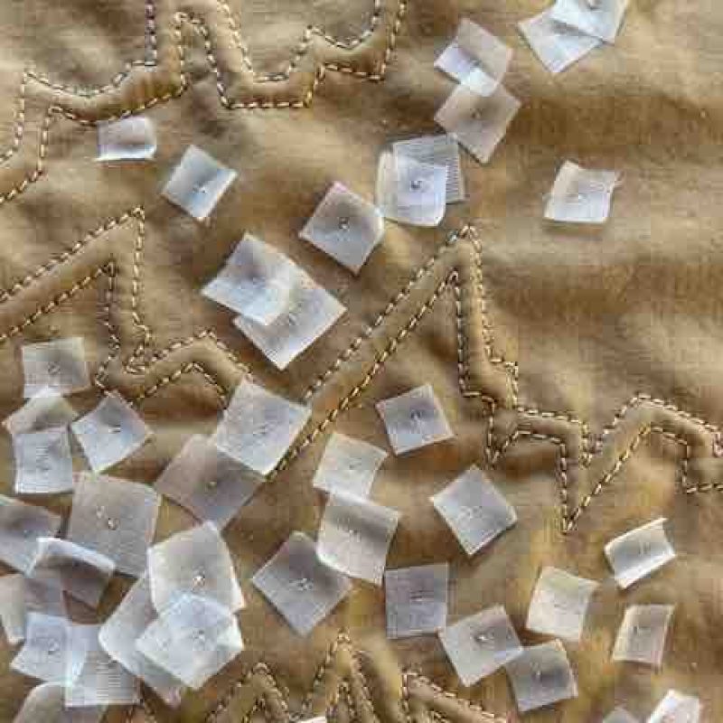 Small white squares of fabric, loosely attached to a beige background with random zig zagging machine stitching