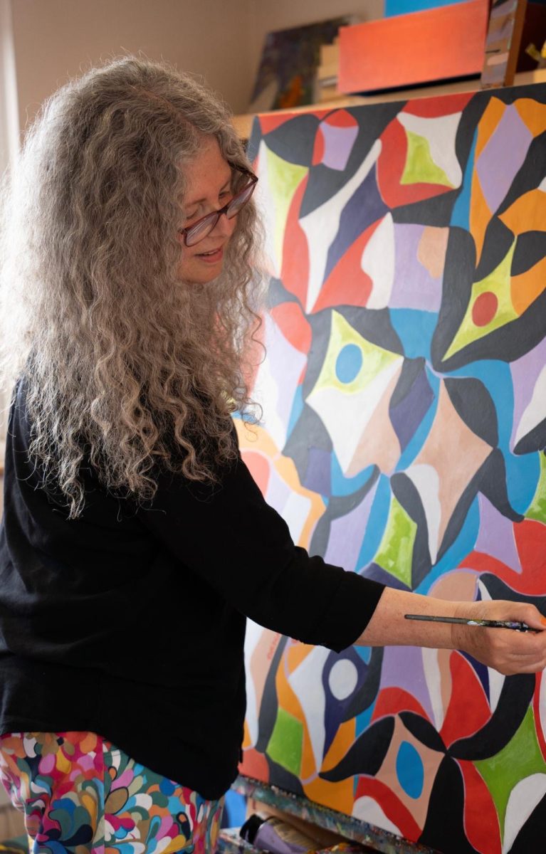 A woman with long grey curly hair holding a brush and painting a large abstract canvas 