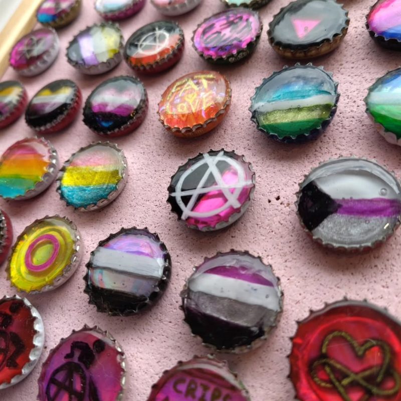 Resin pin badges made from reclaimed bottle caps decorated with colourful designs by the artist Bryony May. Many of the caps feature Pride flags and other symbols related to LGBTQ+ identities.