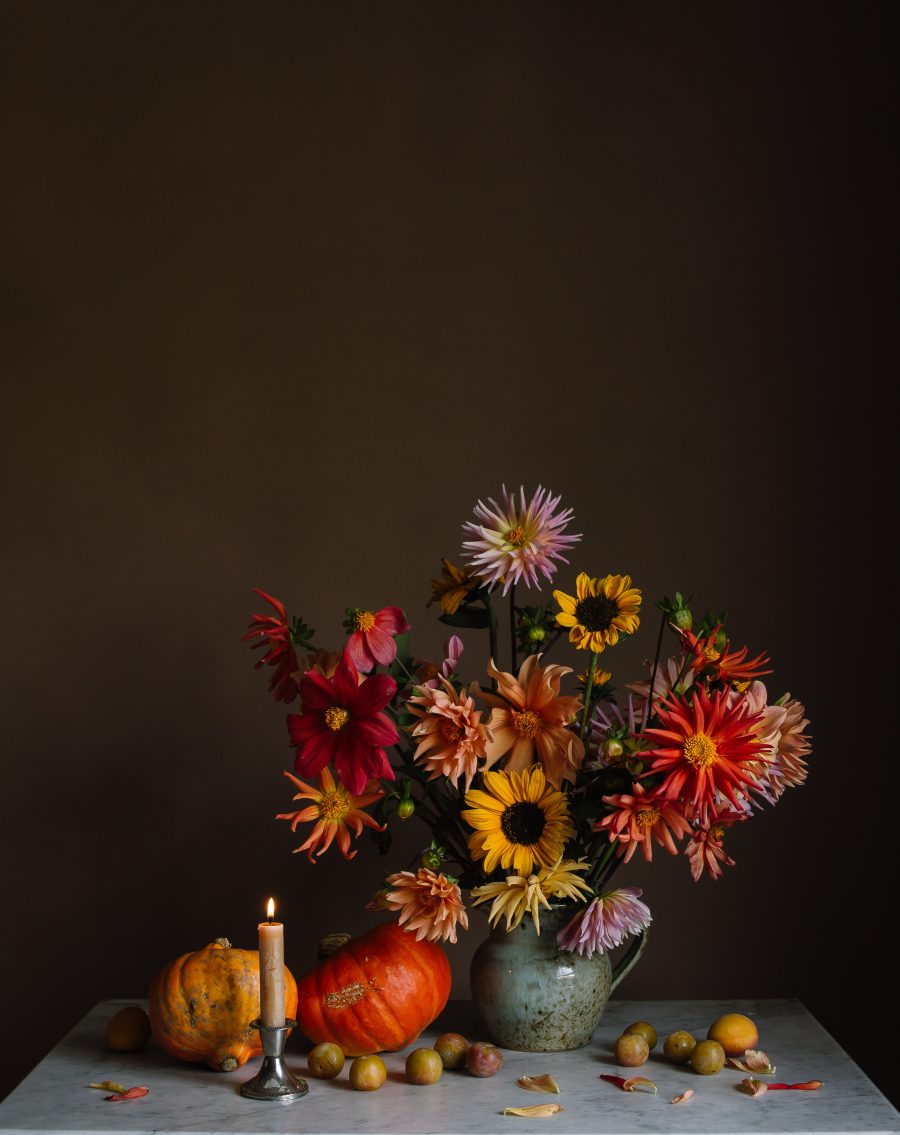 A still-life of dahlias and sunflowers in a jug surrounded by pumpkins and greengages, alongside a lit candle against a dark background