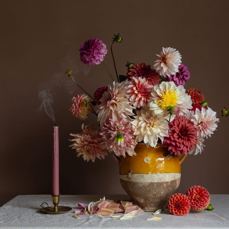 A still-life fine art photograph showing colourful dahlias in a vase alongside a smoking candle against a dark background