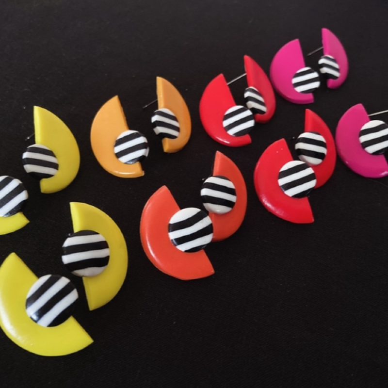 8 pairs of colourful geometric earrings made with semi circles in various warm colours contrasting with a circle of black and white stripes made with polymer clay, displayed on a black background.
