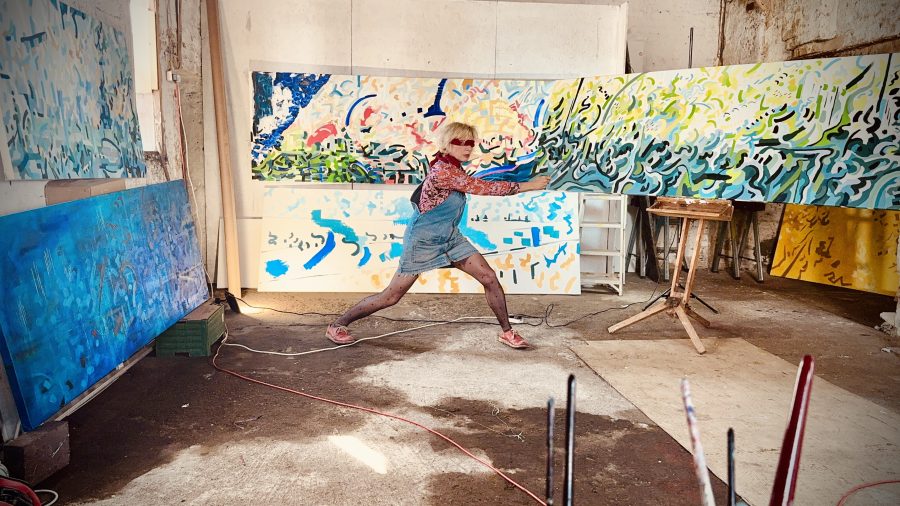 This shows an artist blindfolded, painting her work in a very large studio. There are three colourful canvasses on a huge scale 