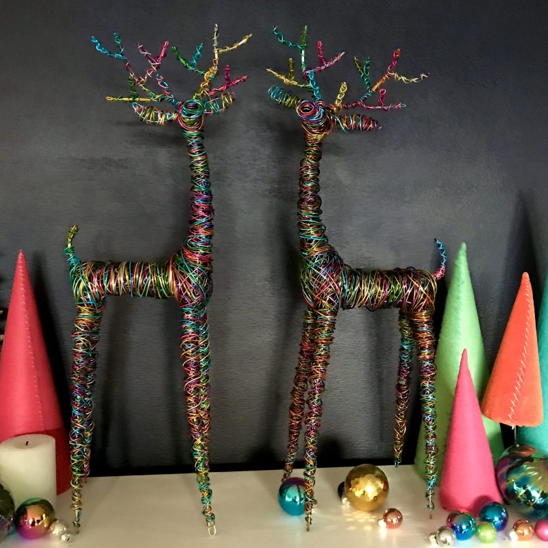 A pair of multicoloured wire reindeer sculptures with elegant elongated proportions and legs that taper to a point, they are standing on a mantelpiece with other colourful decorations around them.