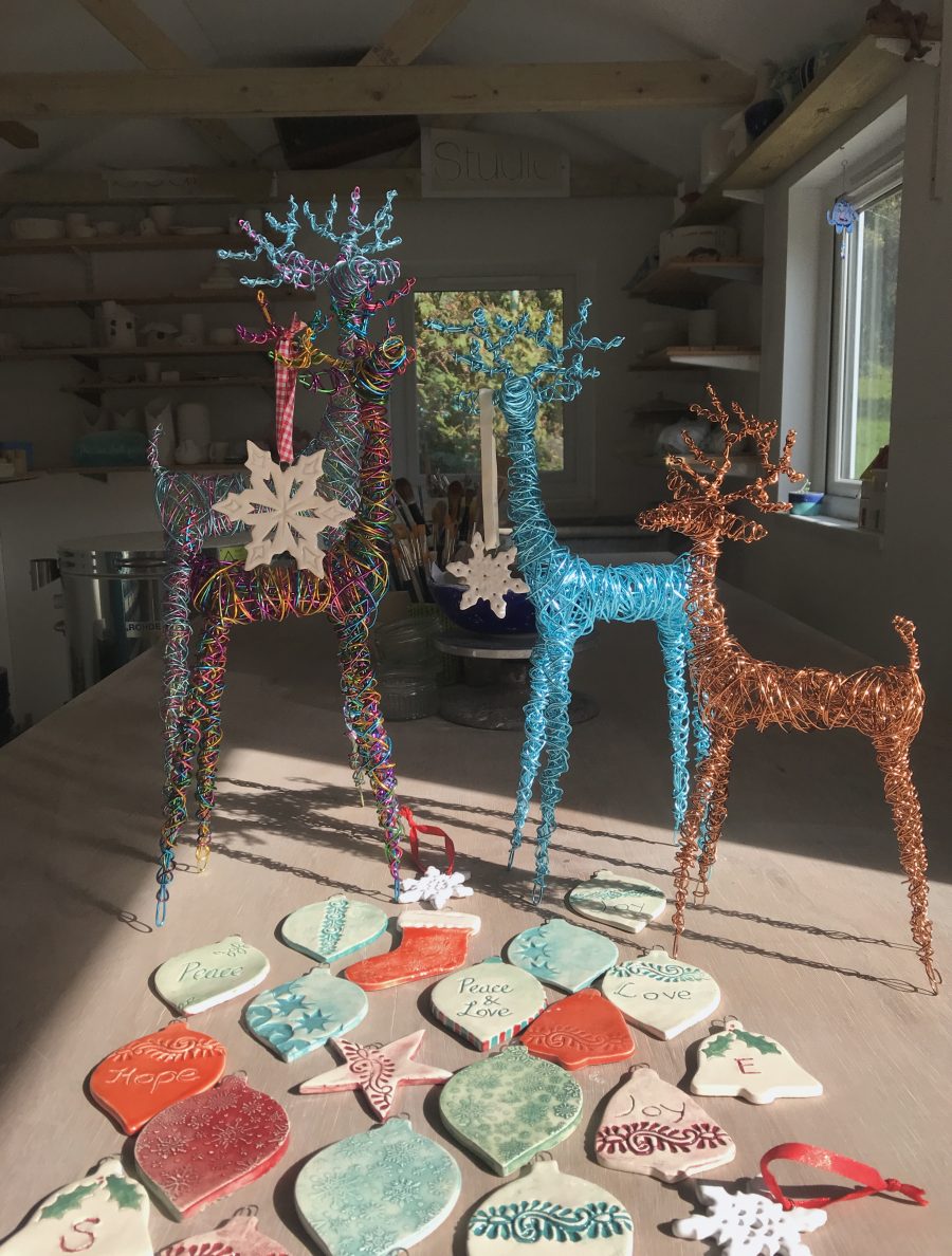 The interior of a small ceramics studio with some elegant  colourful wire reindeer sculptures in the foreground on the worktable and some flat bauble and other shaped ceramic tree decorations some with the words, 'peace', 'love' and 'hope' on them, hanging from their antlers and on the table in front of them.