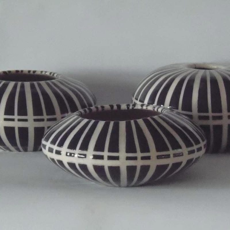 A set of three ceramic vases. They are round and squat shaped, bulging in the middle, decorated with vertical white lines and one horizontal white line around the widest point on a black background. There are two similar vases in the background.