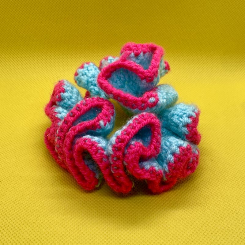 A crocheted hair accessory in light blue and bold pink, photographed against a bold yellow background.