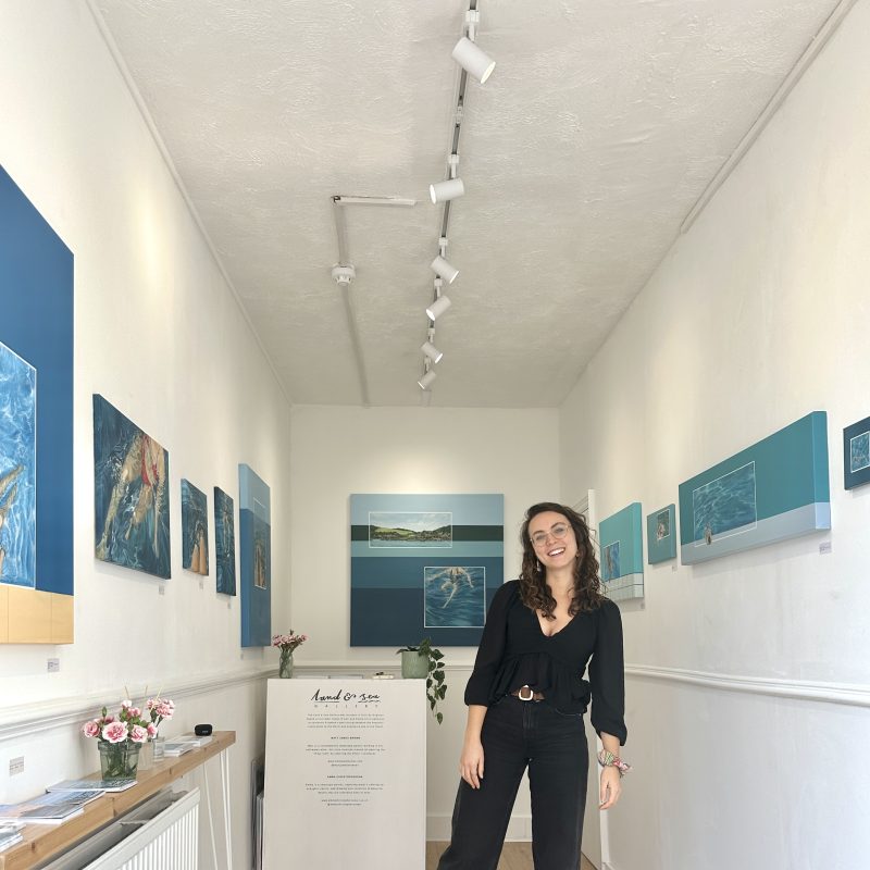 Emma Christopherson in the gallery with paintings of figures in the water 