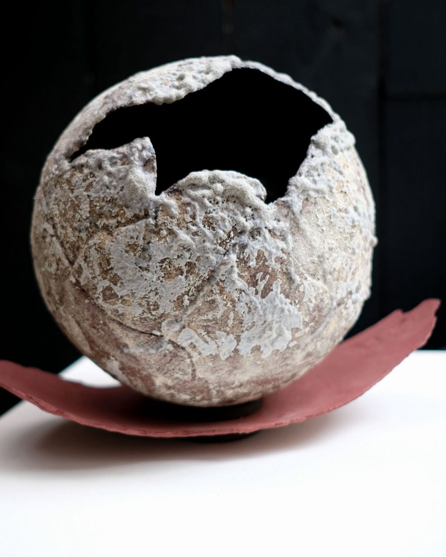 Description for sight impaired: A spherical ceramic ball made of sections of clay, with mouth like opening, flakey white glaze and seated on a red clay curved base.