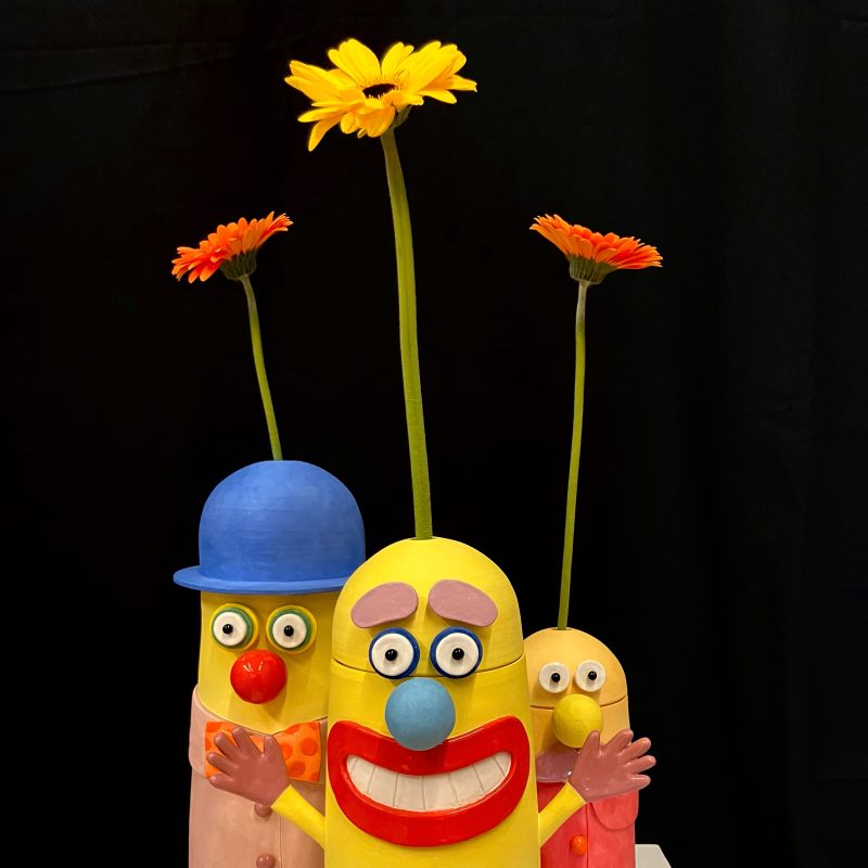 Three ceramic vases all with faces and lids sit on a plinth, each with a single flower stem placed inside.