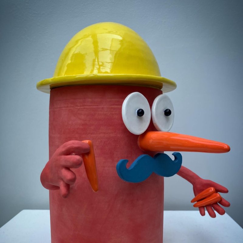 A red vase with a yellow glazed hat, featuring a face with a carrot nose and dark blue moustache; two arms extend from the vase, holding carrots.