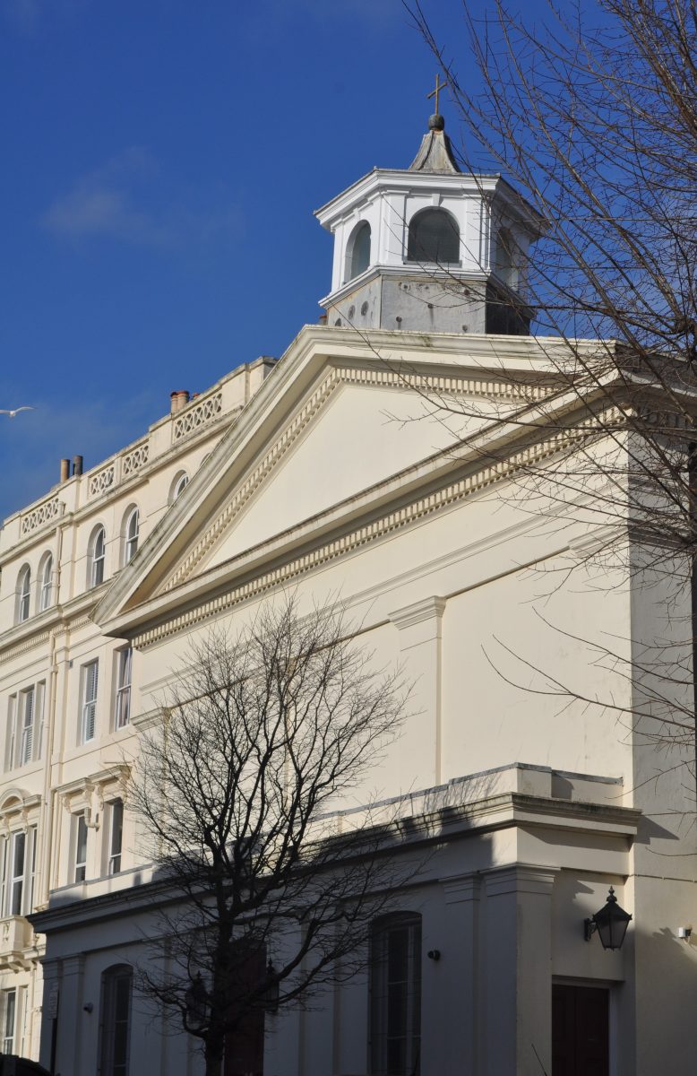 The image shows St Stephen’s Hall in Brighton, a large pale cream building in a classical Georgian style. The front has a wide triangular pediment, like a Greek temple, with simple decorative moulding. Rising from the centre of the roof is a small square tower with arched openings, topped by a rounded dome and a metal cross. The building’s walls are smooth and light-coloured, catching bright sunlight. Shadows from nearby trees fall across the lower part of the structure. In front of the building, leafless tree branches spread upward, suggesting it is winter or early spring. The sky behind the building is a deep, clear blue with no visible clouds, creating a strong contrast with the light-coloured façade. No people or vehicles are visible, and the scene feels quiet and still. 