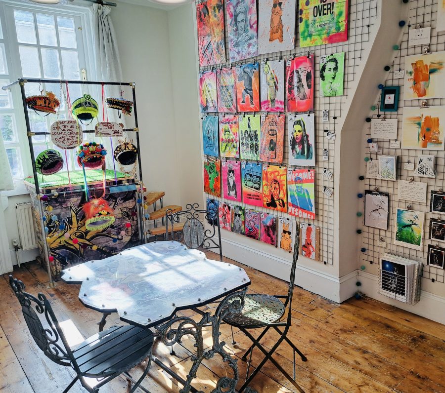 A bright photo from last years exhibition of half of the gallery space with neon pieces of printed art and brightly coloured artistic Hats with a table in the middle of the room.