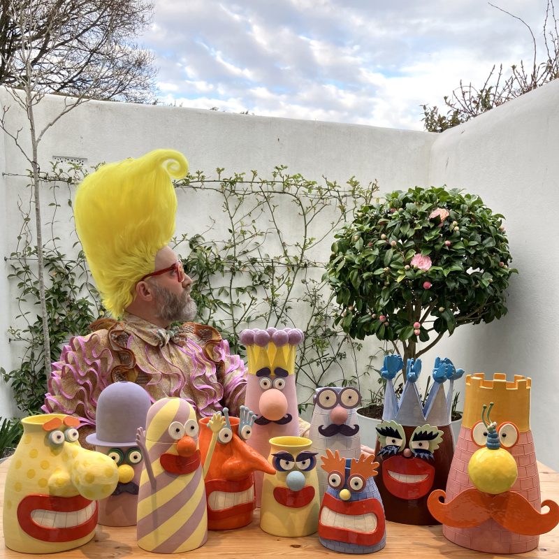 The artist Boogaloo Stu, with bright yellow quiff, over-sized sunglasses and frilled costume, sits beside a selection of his ceramic art placed on a tabletop.
