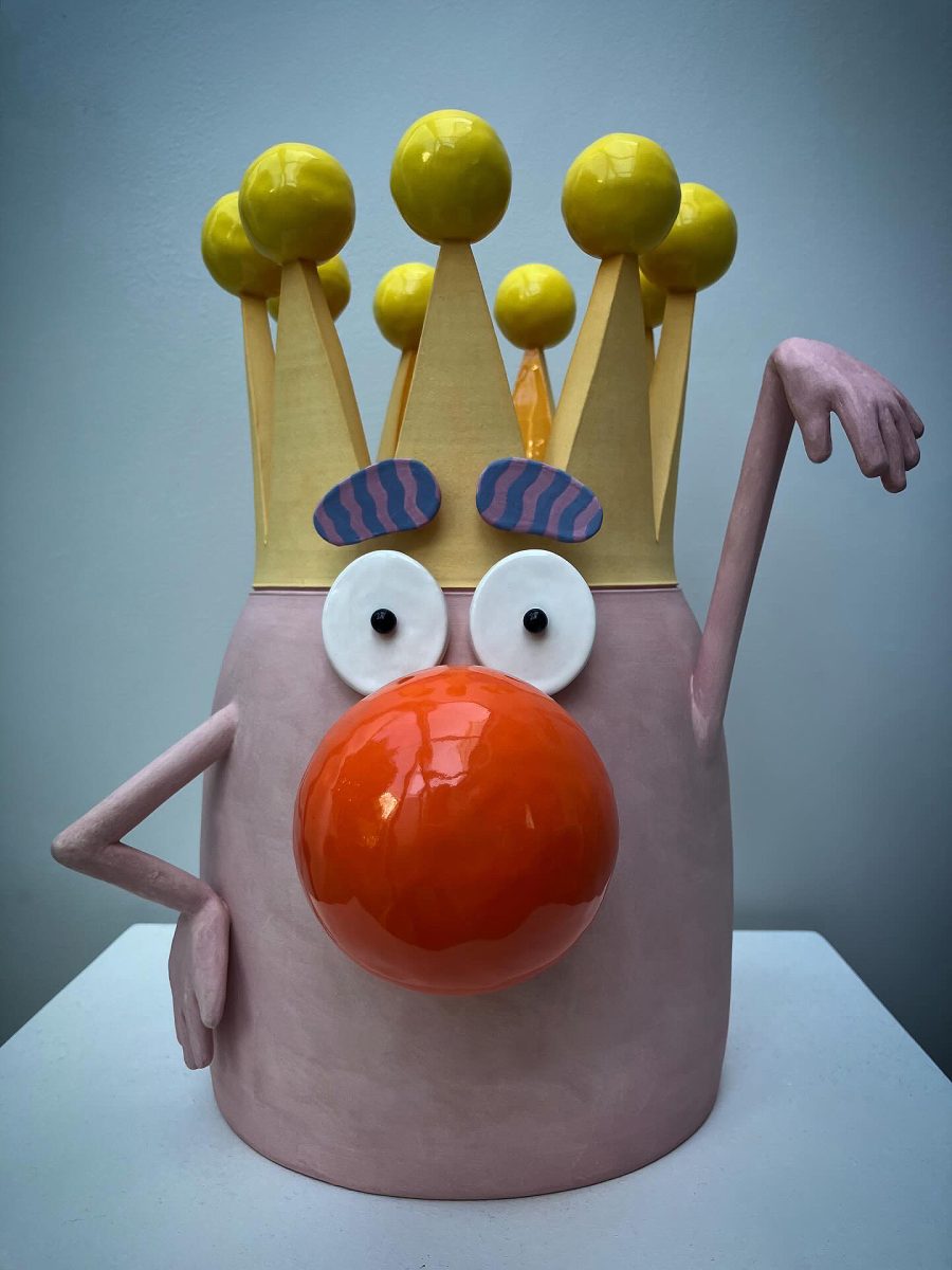 A pink ceramic vase with a gold and yellow crown, features a face with a bright orange nose with arms at either side gesturing flamboyantly.