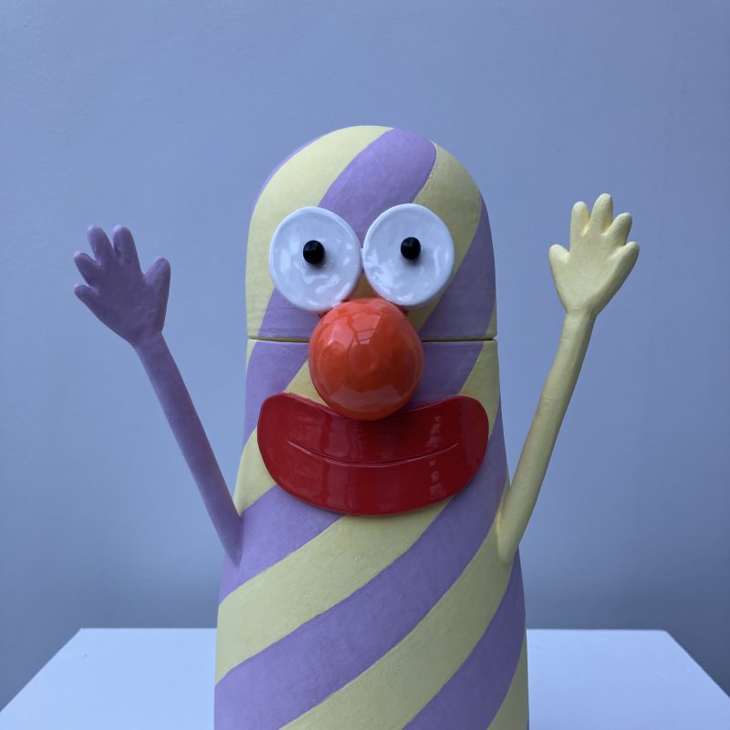 A stripy pink and yellow vase with lid, two arms aloft and a face with orange nose and yellow lips.
