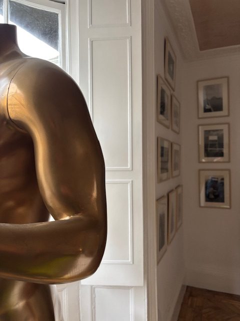 The image shows a corner of the gallery with some framed photographs on one wall and the edge of the gold man statue, which the gallery is named after.