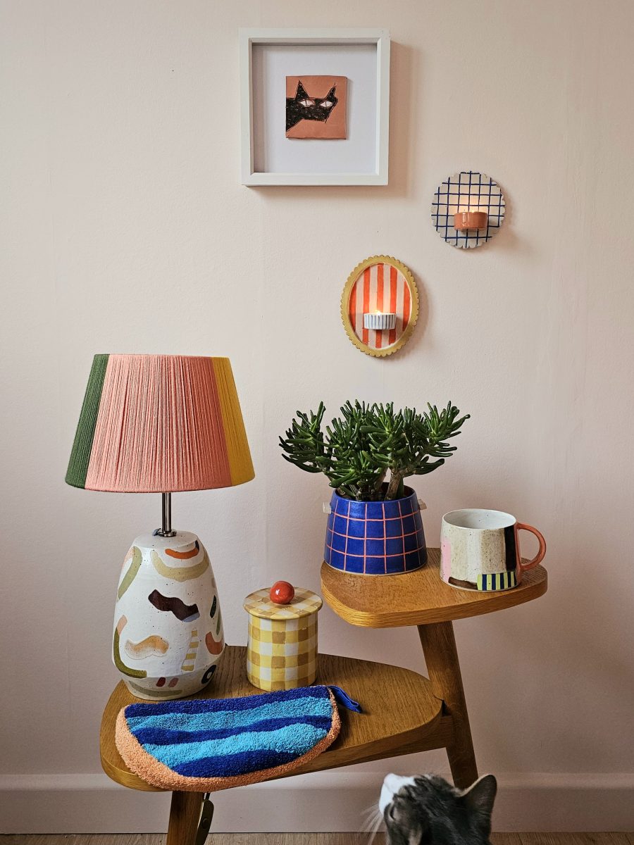 A selection of colourful ceramic pieces are arranged on a table, a print of a cat and 2 ceramic sconces hang on the wall behind