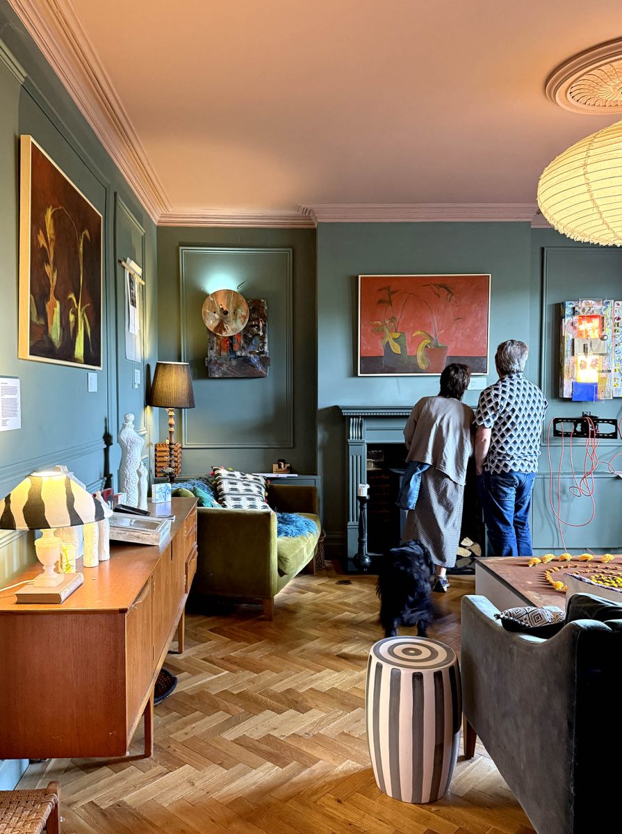 Artworks hang on the wall in a richly coloured interior with parquet floors and a pink ceiling. Two people look at the artworks as a black dog walks past