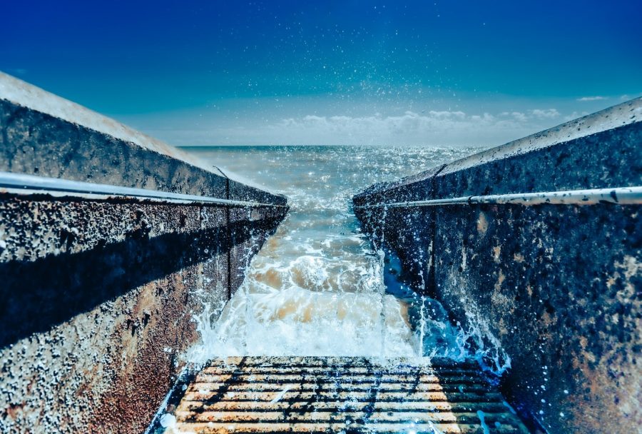 A dramatic seascape viewed from a concrete stairway, where foamy waves crash upward between weathered walls under a bright blue sky.