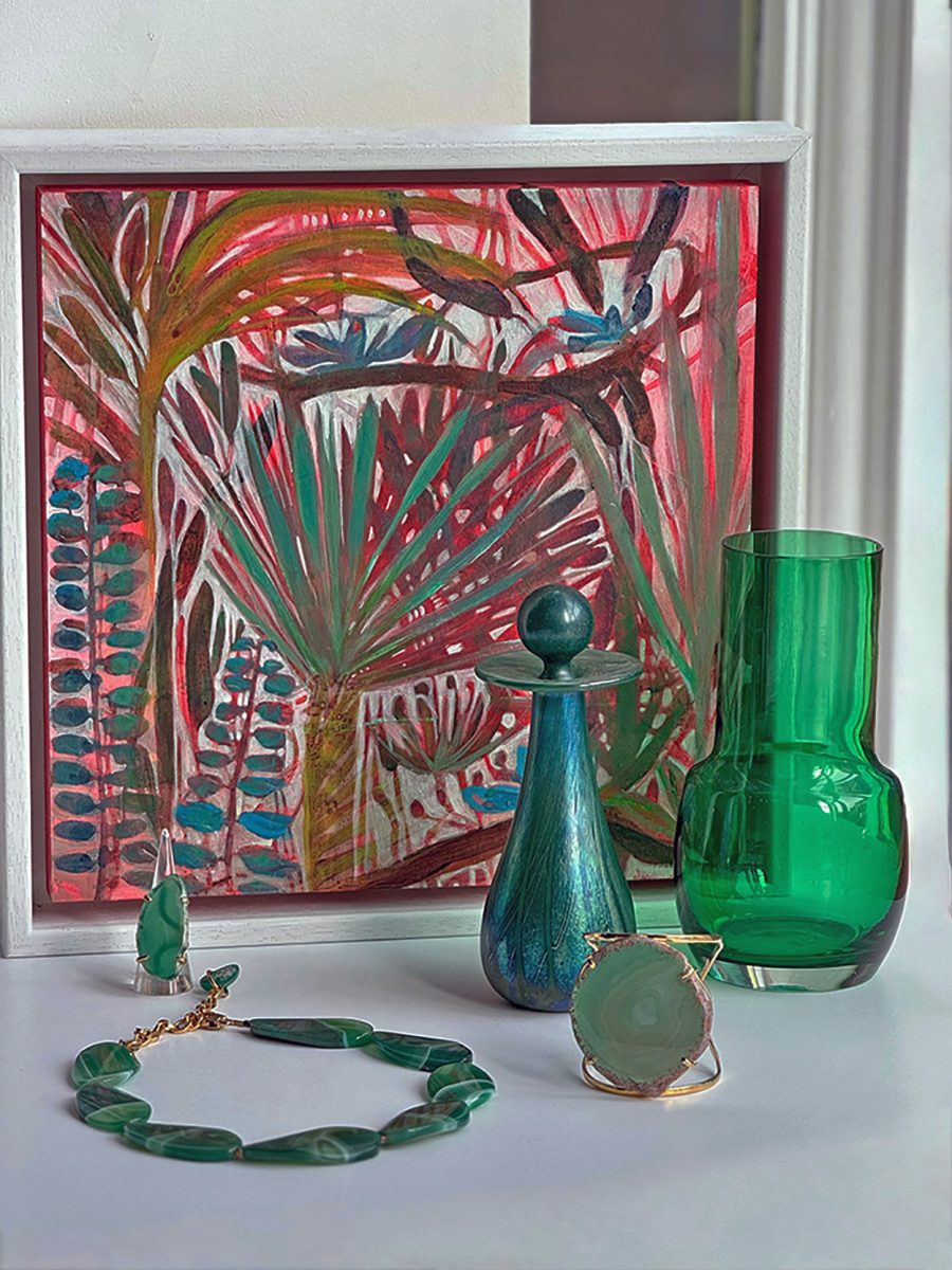 A brightly coloured white framed painting of Jungle plants in reds and greens. Partially covered by a transparent deep green mid century glass vase and an opaque perfume bottle with various jewellery pieces made of  emi precious stones.