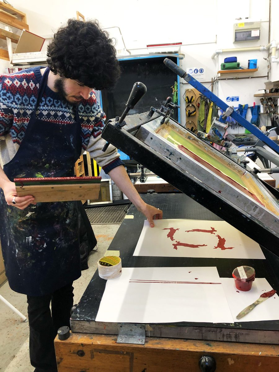 Young man standing in front of a vacuum screenprinting bed holding a squeegee and looking at his recently screenprinted work.