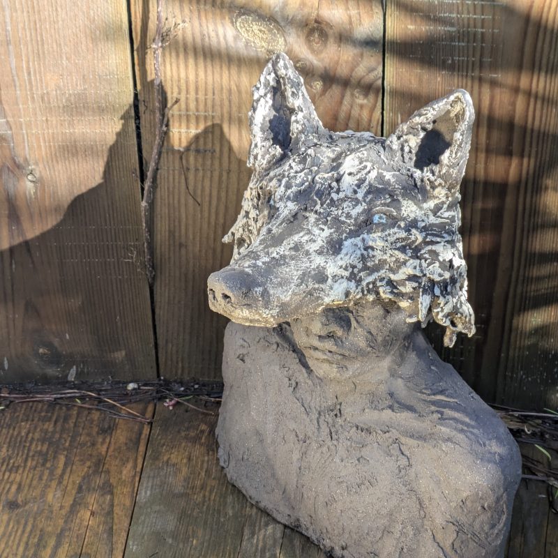 Hand built clay sculpture showing a human head with an animal head covering it. 