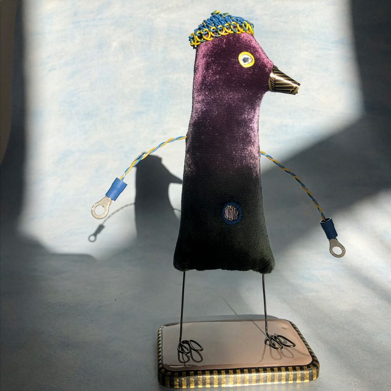 Sculptural two- legged figure with wire legs and arms, and bird-like head. Green and purple velvet body and wire hat.