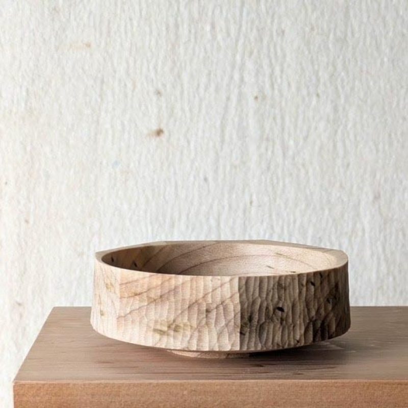 A light‑colored wooden bowl with a textured, hand‑carved surface sits on top of a simple wooden table. The table has clean, minimal lines and angled legs. The background is a plain, off‑white wall with a slightly rough texture. If you’d like, I can also create a shorter version, a longer descriptive version, or alt text optimized for accessibility guidelines (WCAG).