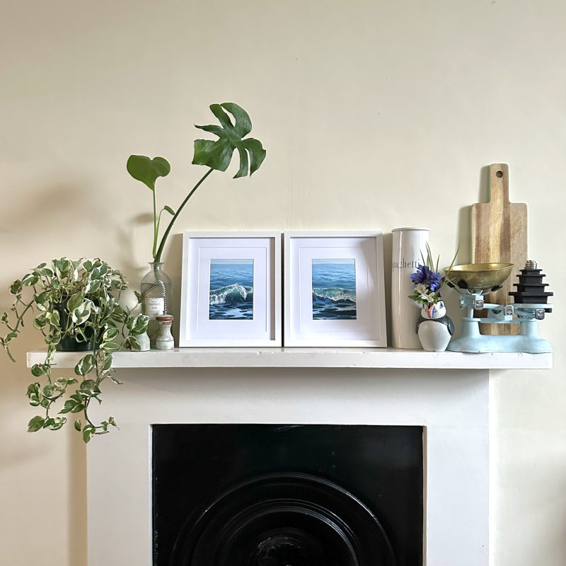 wo framed prints of waves sit on a mantelpiece surrounded by plants and vintage kitchen items. The two waves line up across the two prints. 