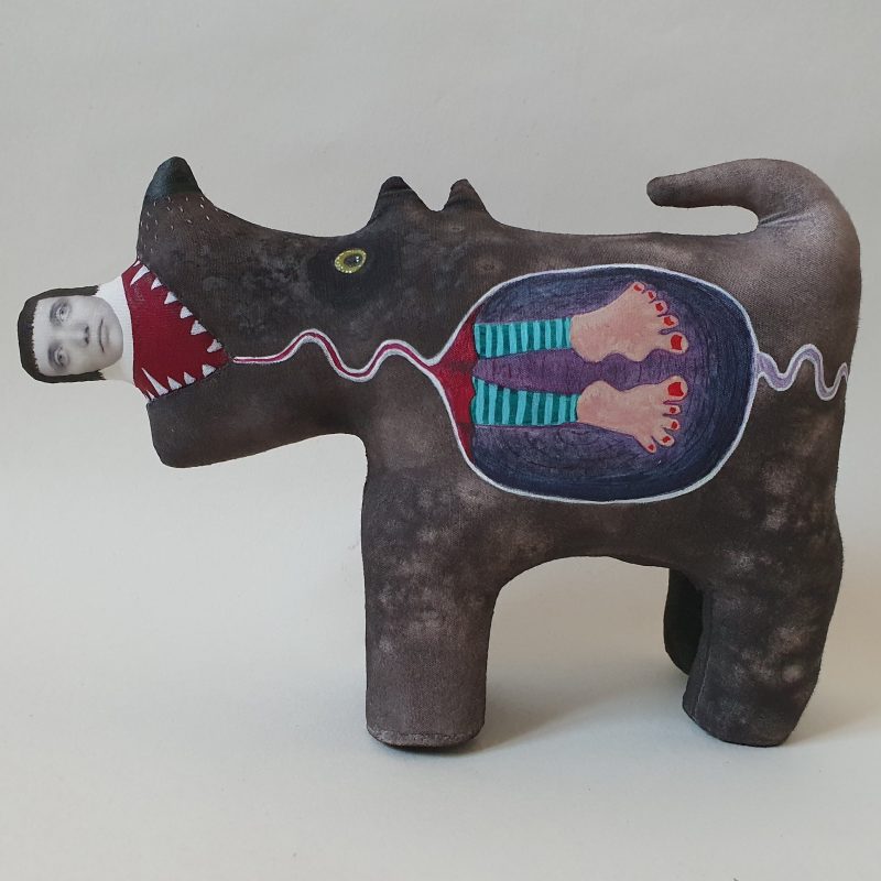 Soft sculpture depicting a brown dog eating its owner, who is lying rigid in the dog's mouth. The side of the dog is painted to show the inside of the dog with the rigid owners feet in the dog's stomach.