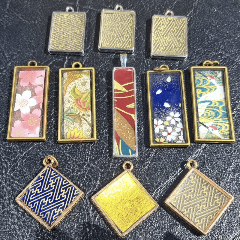 A collection of decorative pendants with various patterns and motifs is displayed on a dark surface. The pendants feature designs like cherry blossoms, cranes, and geometric patterns.