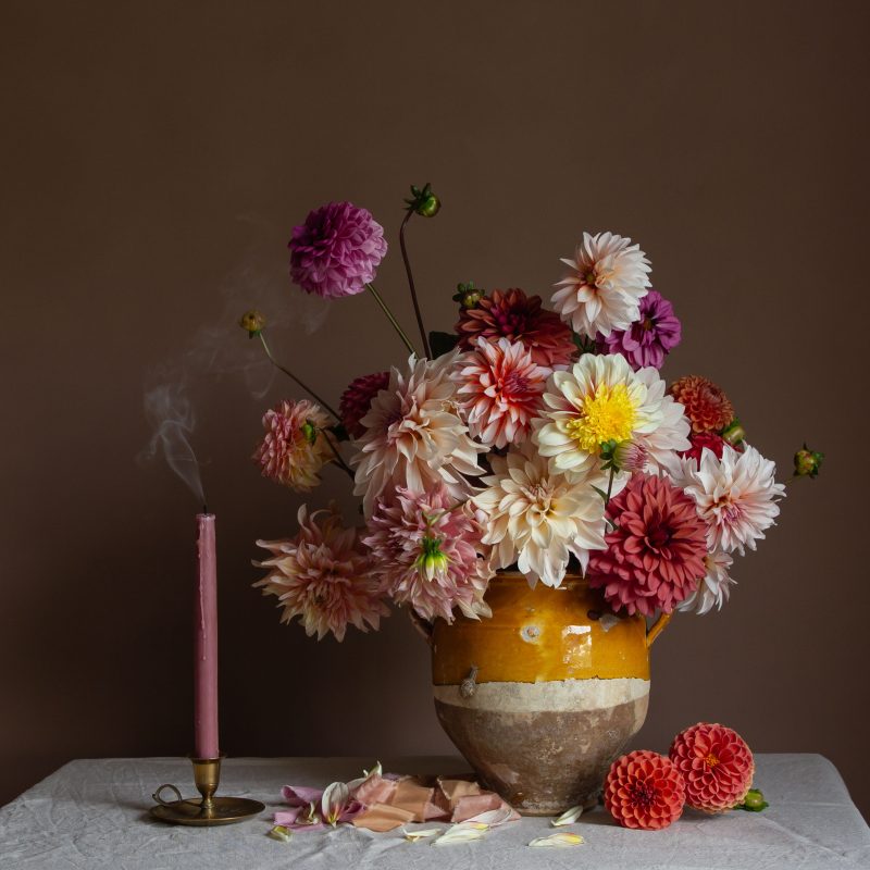 A still life fine art photographer showing dahlias in an old yellow vessel alongside a smoking candle
