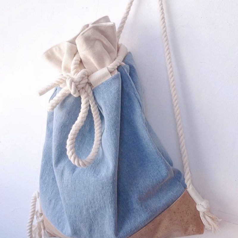 A drawstring bag in natural cotton twill and main part in a light denim fabric and light coloured cork bottom with a thick cotton cord drawstring top and handles to carry on your back