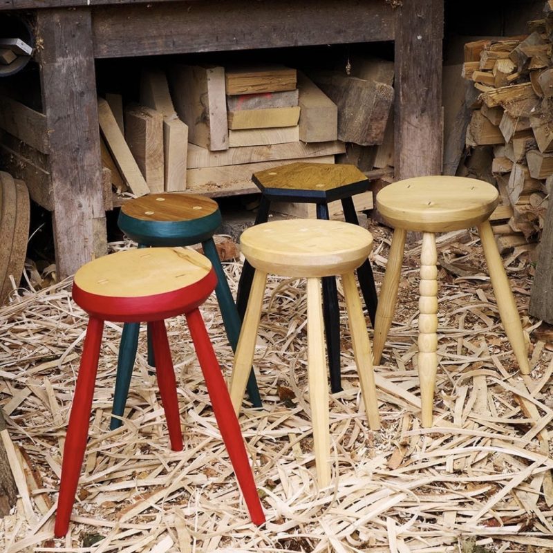 Hand Made 3 legged Wooden stools sitting on sawdust covered floor. Two stools are natural wood colour and one stool has red painted edges and leg  details.