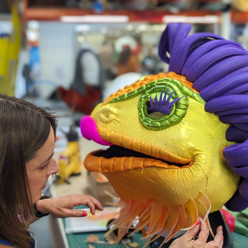 A woman is attaching pieces of fabric to a large colourful dragon costume head.
