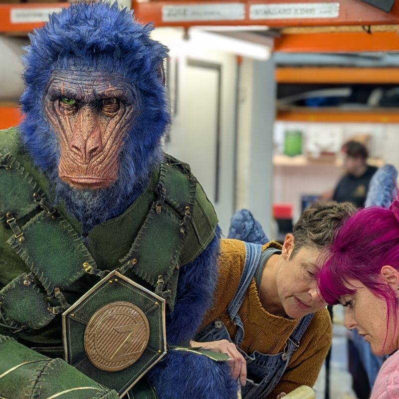 Two makers are carefully glueing on fur to a scale model of Chistery, the winged monkey from the Wicked For Good movie.