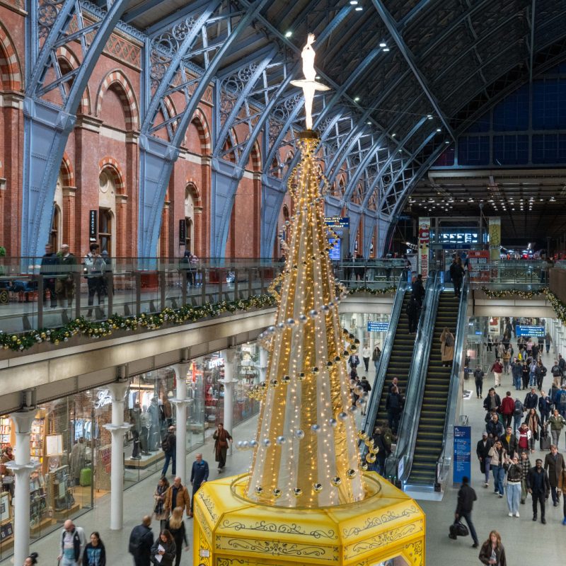 A sparkling yellow Christmas tree in the shape of a giant music box sits in the middle of a busy railway concourse. Silver and gold baubles wind up a striped fabric cone with an illuminated ballerina at the top. 
