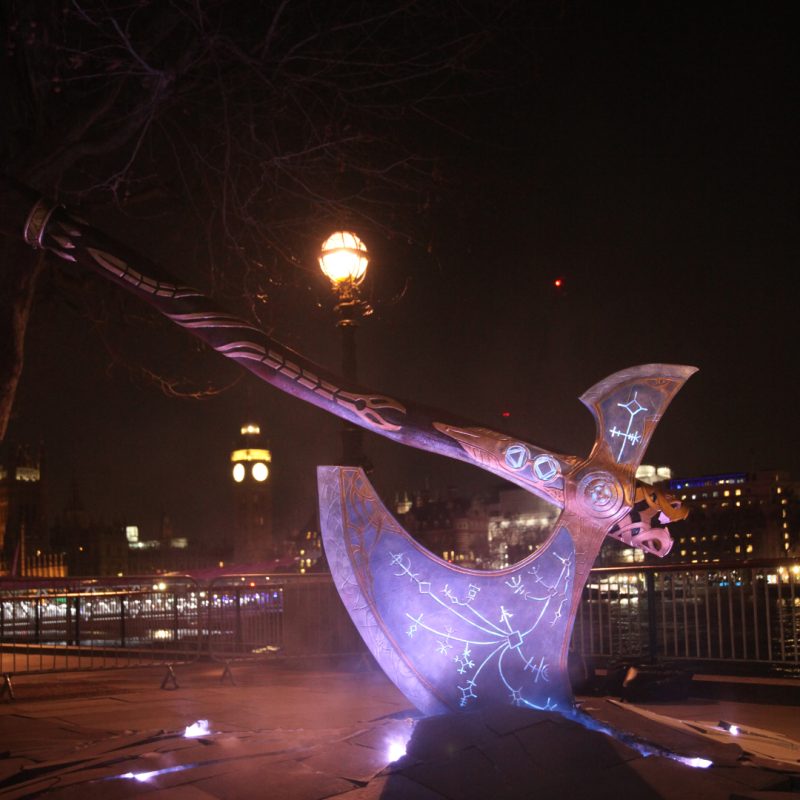 A gigantic axe is sticking out of cracked paving stones beside the river Thames. Smoke is coming out of the ground and the axe is covered in illuminated marks and symbols.
