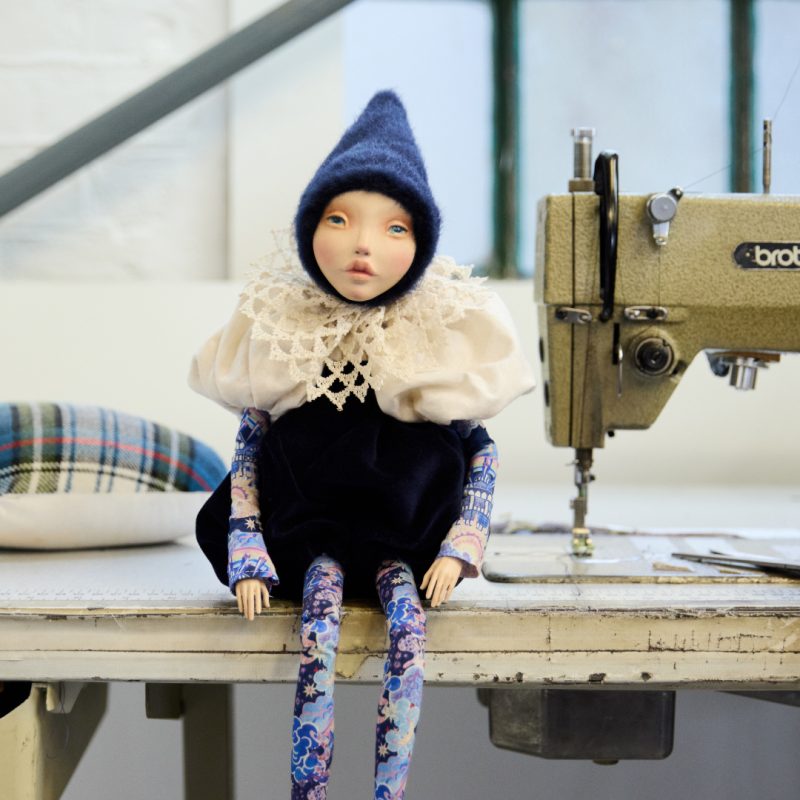 A small, delicate elf doll sits on a workbench beside a sewing machine and pin cushion. The elf is dressed in a pointed blue felt hat, puffy cream collar and old fashioned button boots. Its legs and arms are covered in Liberty print fabric