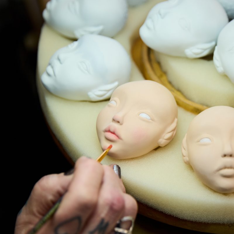 Multiple blank doll faces are laid out on a bench. An artist is using a tiny brush to hand paint one of the faces.