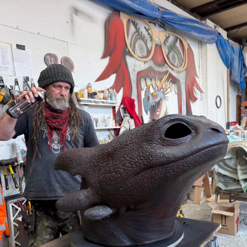A man is standing in a colourful creative workshop space. He is spray painting a large dragon head model. There are tools and artistic equipment on the wall behind him.