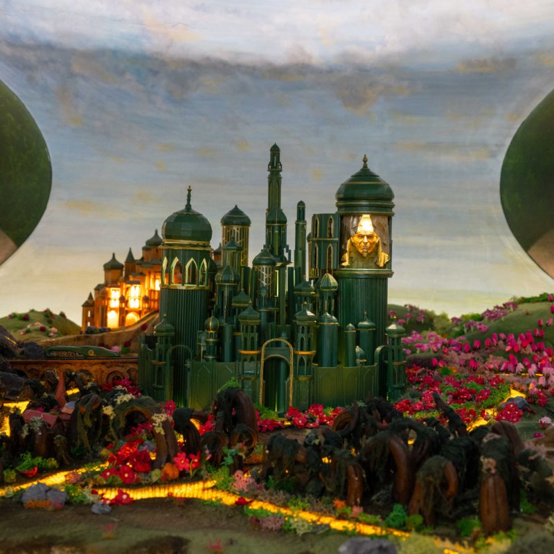 A tiny model diorama of the Emerald City from the movie Wicked, handmade by the team at Plunge Creations. The yellow brick road is illuminated and leads through fields towards a cityscape of tiny lit up turrets.