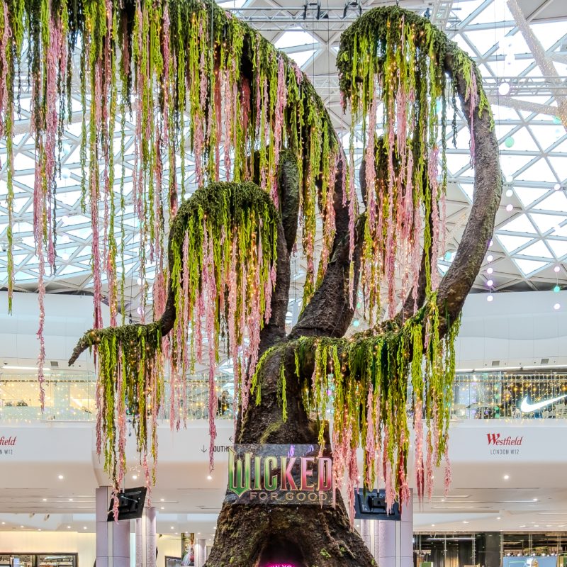 A 12 metre high hand made tree inside a shopping centre, inspired by the film Wicked For Good. The curved branches are draped in pink and green foliage. There's a seat in the base of the trunk with a pink and green neon sign behind for photo ops.